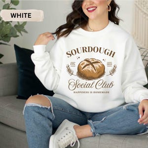 May include: White sweatshirt with the text "SOURDOUGH Social Club" and a graphic of a loaf of bread. The text "Happiness is homemade" is below the club name. The sweatshirt is worn with blue jeans and white sneakers.