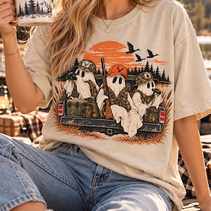 May include: A beige short-sleeve t-shirt featuring a graphic of three ghosts sitting in the back of a truck. The ghosts wear hunting hats and camouflage gear, with one holding a rifle and others using binoculars. An orange sun and flying ducks are visible.