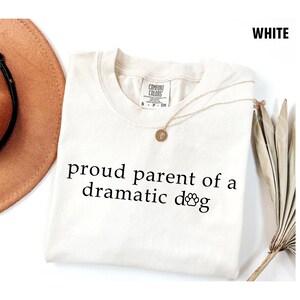 May include: Cream-colored t-shirt with the text "proud parent of a dramatic dog" in black. A gold necklace with a pendant rests on the shirt. The shirt is labeled "Comfort Colors" and is a size small.