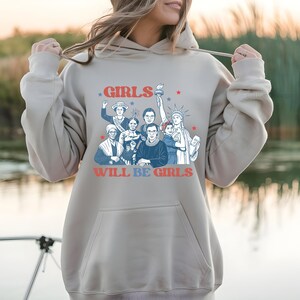 May include: A light beige hoodie with a graphic featuring a group of women and the text "GIRLS WILL BE GIRLS" in red and blue. The design includes illustrations of historical figures and the Statue of Liberty.