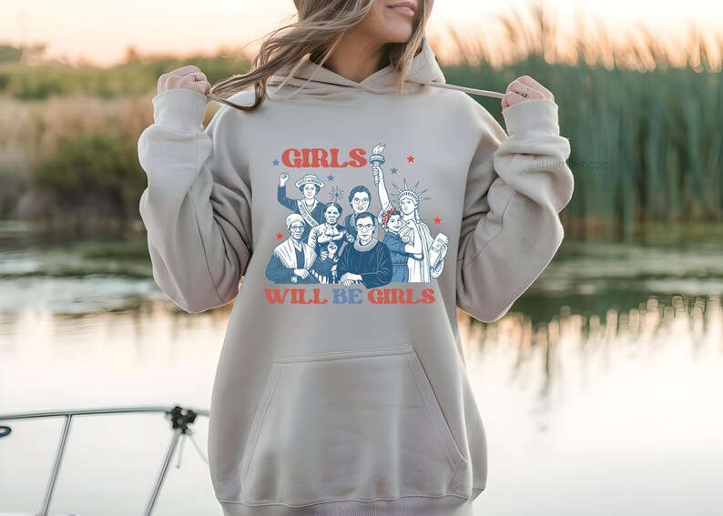 May include: A light beige hoodie with a graphic featuring a group of women and the text "GIRLS WILL BE GIRLS" in red and blue. The design includes illustrations of historical figures and the Statue of Liberty.