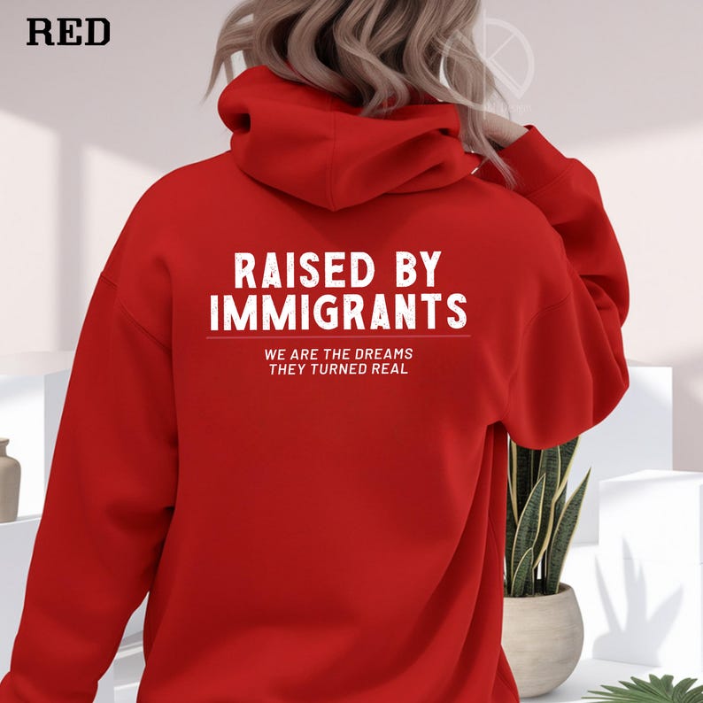 May include: A red hoodie with the text "RAISED BY IMMIGRANTS" in white, with the tagline "WE ARE THE DREAMS THEY TURNED REAL". The word "RED" is in the top left corner. The hoodie has a hood and long sleeves.