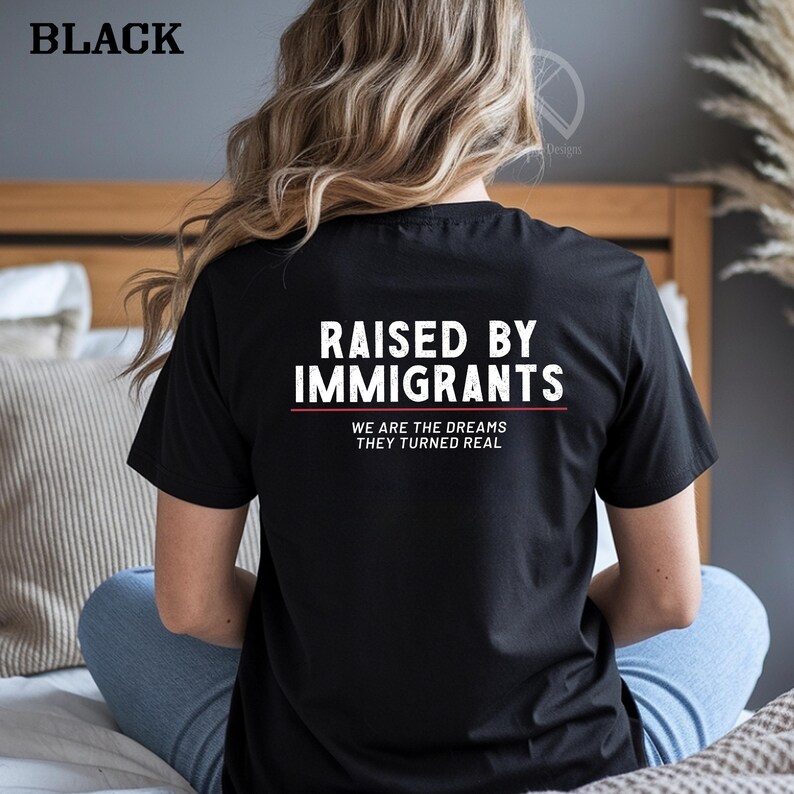 May include: Black t-shirt with the text "RAISED BY IMMIGRANTS" in white, with a red line underneath. Below the line, the text reads "WE ARE THE DREAMS THEY TURNED REAL". The word "BLACK" is in the top left corner.
