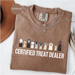 May include: Espresso-colored t-shirt with the text "CERTIFIED TREAT DEALER" in white. The shirt features a row of colorful paw prints. A silver necklace with a pendant is visible.