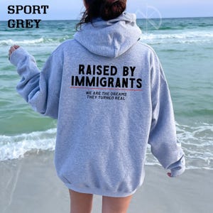 May include: Sport grey hoodie with the text "RAISED BY IMMIGRANTS" in black block letters above a red line. Below the line, the text reads "WE ARE THE DREAMS THEY TURNED REAL" in a smaller font. The hoodie is shown from the back.