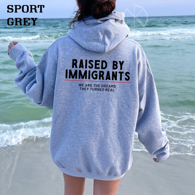 May include: Sport grey hoodie with the text "RAISED BY IMMIGRANTS" in black block letters above a red line. Below the line, the text reads "WE ARE THE DREAMS THEY TURNED REAL" in a smaller font. The hoodie is shown from the back.