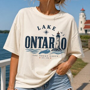 May include: A cream-coloured short-sleeve t-shirt featuring a graphic print that reads Lake Ontario, The Great Lakes, and Est. 1792. The design includes a compass rose, a lighthouse, birds, and blue waves in dark blue and light blue ink.