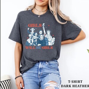 May include: Dark heather gray t-shirt featuring a graphic of historical and iconic women with the text "GIRLS WILL BE GIRLS" in red and blue. The shirt is a crew neck and has short sleeves. The shirt is paired with blue jeans.