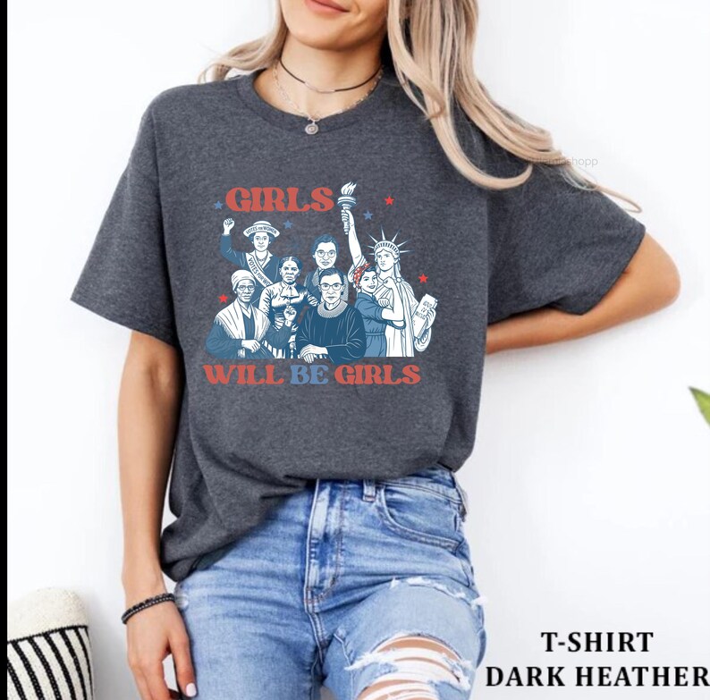 May include: Dark heather gray t-shirt featuring a graphic of historical and iconic women with the text "GIRLS WILL BE GIRLS" in red and blue. The shirt is a crew neck and has short sleeves. The shirt is paired with blue jeans.