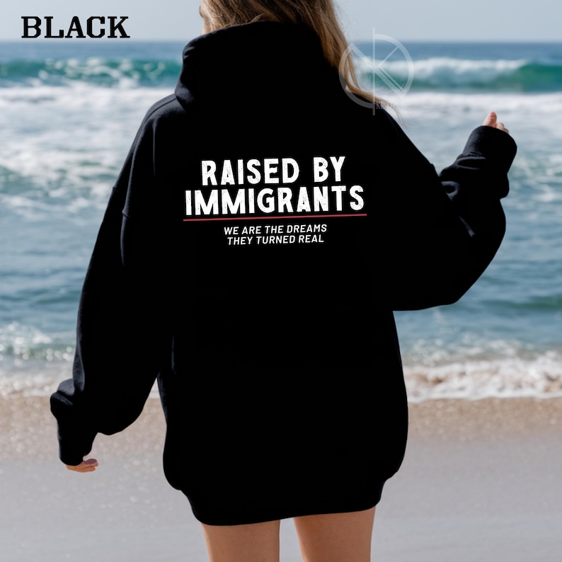 May include: Black hoodie with the text "RAISED BY IMMIGRANTS" in white, with the words "WE ARE THE DREAMS THEY TURNED REAL" below. The word "BLACK" is at the top left corner of the image. The background is a beach with ocean waves.