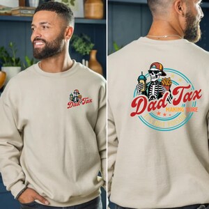 以下が含まれることがあります： 前面と背面にグラフィックが施されたベージュのクルーネックのトレーナー。前面のグラフィックは、帽子をかぶった骸骨で「Dad Tax」と書かれています。背面のグラフィックは、飲み物とフライドポテトを持っている骸骨で「Dad Tax」と書かれています。