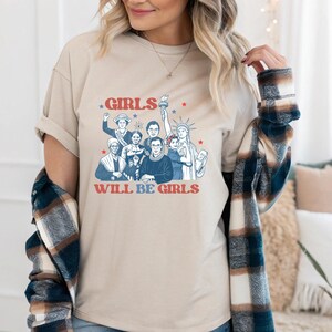 May include: Beige t-shirt featuring a graphic of diverse women with the text "GIRLS WILL BE GIRLS" in red and blue. The design includes illustrations of historical figures and symbols of freedom. The shirt has short sleeves.