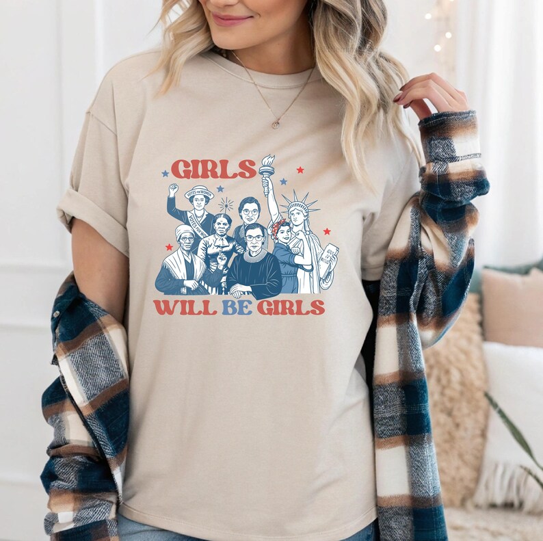 May include: Beige t-shirt featuring a graphic of diverse women with the text "GIRLS WILL BE GIRLS" in red and blue. The design includes illustrations of historical figures and symbols of freedom. The shirt has short sleeves.