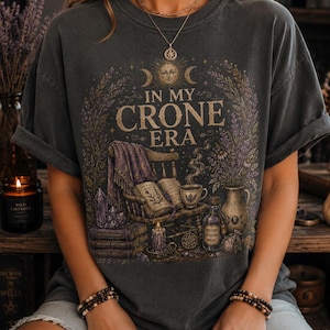 May include: A charcoal gray t-shirt featuring a graphic print with the text IN MY CRONE ERA. The design includes a sun and moon, a wooden chair with a purple blanket, an open book, a steaming cup, a candle, crystals, and a vase of flowers.