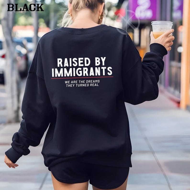 May include: Black sweatshirt with the text "RAISED BY IMMIGRANTS" in white, with a red line underneath. Below the text is the phrase "WE ARE THE DREAMS THEY TURNED REAL" in white. The word "BLACK" is in the top left corner.