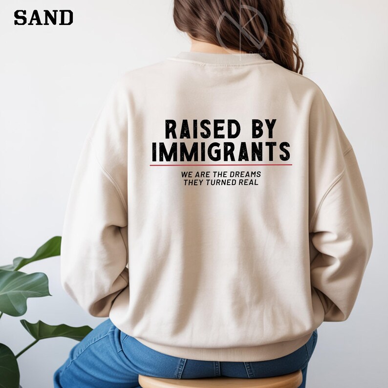 May include: A sand-colored sweatshirt with the text "RAISED BY IMMIGRANTS" in black, with a red line underneath. Below the text is "WE ARE THE DREAMS THEY TURNED REAL" in a smaller font. The word "SAND" is in the top left corner.