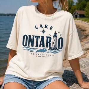 May include: An off-white short-sleeve t-shirt featuring a dark blue graphic print that reads LAKE ONTARIO with a lighthouse, a compass rose, and waves. Below the graphic, the text reads THE GREAT LAKES EST. 1792 with two small red stars.