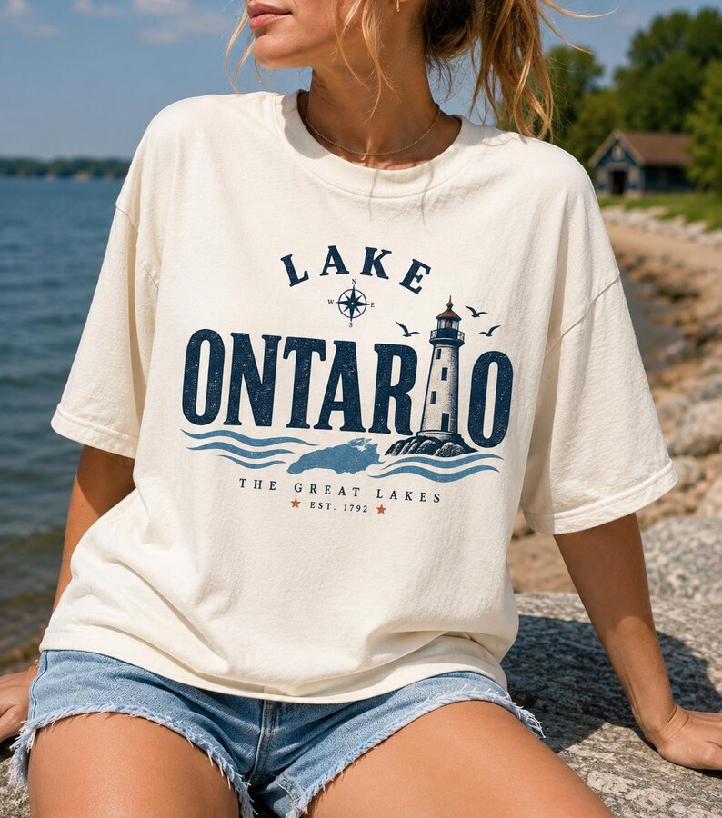 May include: An off-white short-sleeve t-shirt featuring a dark blue graphic print that reads LAKE ONTARIO with a lighthouse, a compass rose, and waves. Below the graphic, the text reads THE GREAT LAKES EST. 1792 with two small red stars.