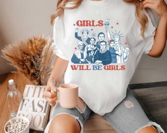 May include: White t-shirt with a graphic featuring illustrations of historical women and the text "GIRLS WILL BE GIRLS" in red and blue. The design includes stars and a torch-bearing figure.