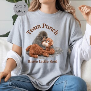 May include: Sport grey t-shirt with the text "Team Punch" and "Brave Little Soul". The shirt features a graphic of two monkeys hugging. One monkey is gray, and the other is brown. The shirt is made of a soft material.