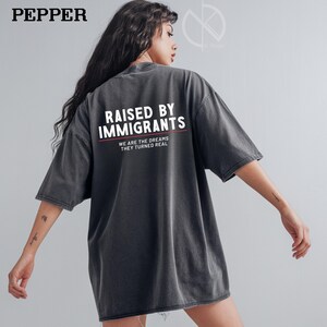 May include: Dark gray oversized t-shirt with the text "RAISED BY IMMIGRANTS" in white, with the words "WE ARE THE DREAMS THEY TURNED REAL" underneath. The shirt has short sleeves and a relaxed fit.