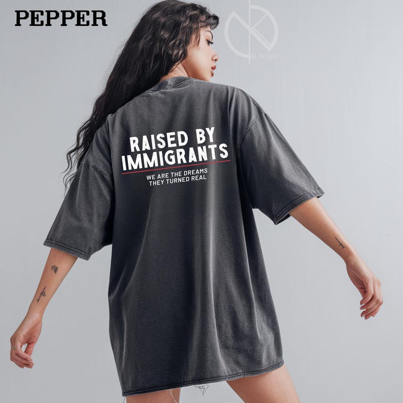 May include: Dark gray oversized t-shirt with the text "RAISED BY IMMIGRANTS" in white, with the words "WE ARE THE DREAMS THEY TURNED REAL" underneath. The shirt has short sleeves and a relaxed fit.