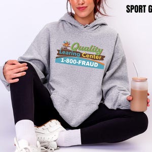 May include: Sport grey hoodie with the text "Quality Learning Center 1-800-FRAUD" in a colorful design. The hoodie is paired with black leggings and white sneakers. A glass with a brown beverage is held in the hand.