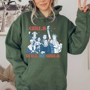 May include: Olive green hoodie with a graphic featuring the text "GIRLS WILL BE GIRLS" in red and blue. The graphic includes illustrations of historical figures and the Statue of Liberty, with stars scattered around the text.
