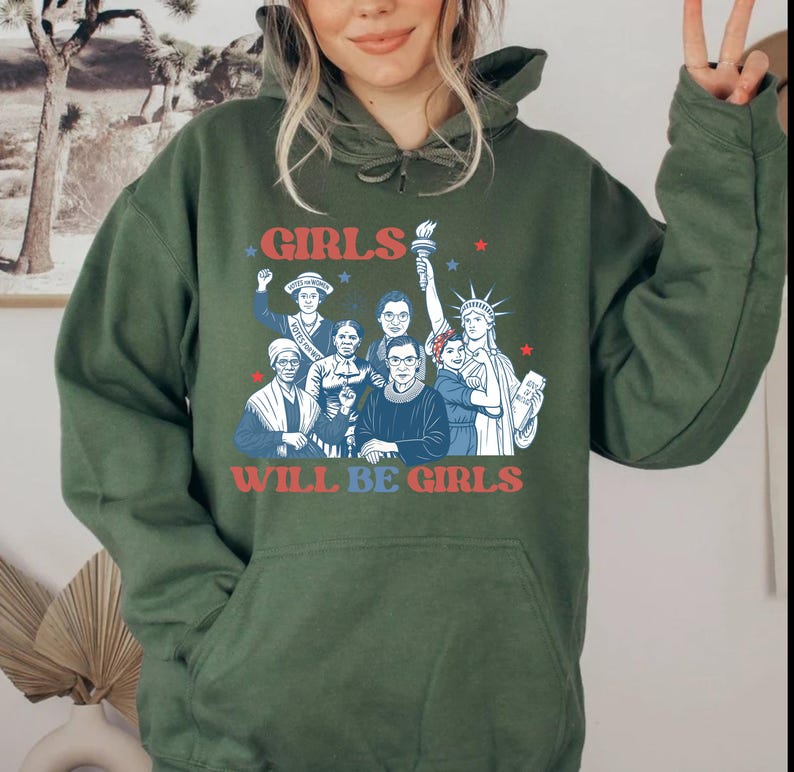May include: Olive green hoodie with a graphic featuring the text "GIRLS WILL BE GIRLS" in red and blue. The graphic includes illustrations of historical figures and the Statue of Liberty, with stars scattered around the text.