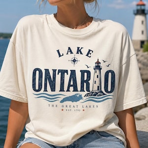 May include: A cream-coloured short-sleeve t-shirt featuring a graphic print that reads Lake Ontario, The Great Lakes, Est. 1792. The design includes a lighthouse, a compass rose, birds, and blue waves, with a map silhouette of the lake.