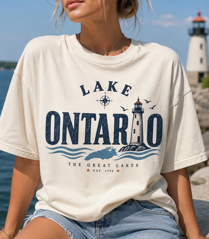 May include: A cream-coloured short-sleeve t-shirt featuring a graphic print that reads Lake Ontario, The Great Lakes, Est. 1792. The design includes a lighthouse, a compass rose, birds, and blue waves, with a map silhouette of the lake.