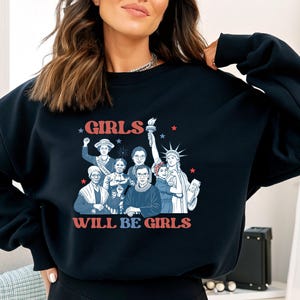 May include: Navy blue sweatshirt with a graphic featuring a group of women and the text "GIRLS WILL BE GIRLS" in red and white. The design includes illustrations of historical figures and the Statue of Liberty.