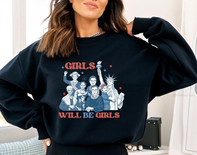 May include: Navy blue sweatshirt with a graphic featuring a group of women and the text "GIRLS WILL BE GIRLS" in red and white. The design includes illustrations of historical figures and the Statue of Liberty.