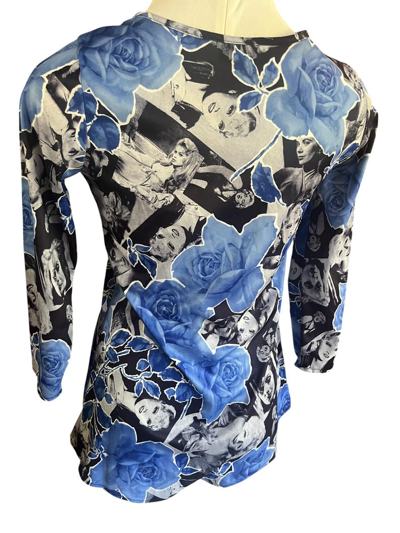 May include: A long-sleeved top featuring a collage of black and white images with large blue rose floral accents. The top has a scoop neck and is made of a lightweight, possibly sheer fabric. The design is repeated across the entire garment.