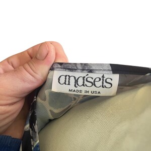 May include: A close-up of a garment tag with the brand name "anasets" in a stylized font. The tag also includes the text "MADE IN USA" in a smaller font. The fabric has a blue, black, and white floral pattern.