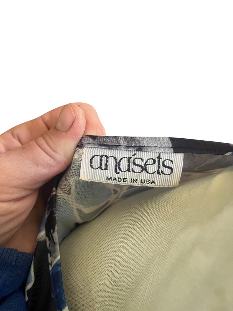 May include: A close-up of a garment tag with the brand name "anasets" in a stylized font. The tag also includes the text "MADE IN USA" in a smaller font. The fabric has a blue, black, and white floral pattern.