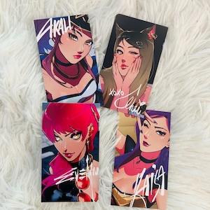 May include: Four art prints featuring stylized illustrations of female characters. Each print showcases a different character with unique hairstyles, outfits, and poses. The prints have the names "Akali", "Ahri", "Seraphine", and "Kai'Sa" written on them.