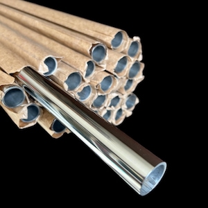 May include: A stack of shiny, silver metal tubes, with a single tube in the foreground. The tubes are cylindrical and have a polished, reflective surface. The tubes are wrapped in brown paper. The background is black.
