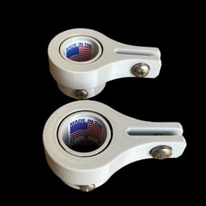 May include: Two white, mechanical components with a circular opening displaying an American flag and the text "MADE IN THE U.S." Each component has a curved arm with a screw. The background is black.
