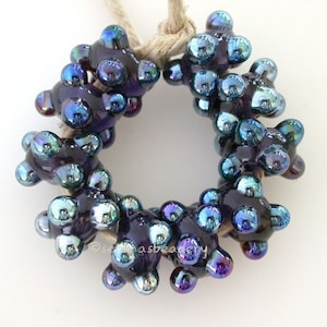Könnte beinhalten: Nahaufnahme einer dunkelvioletten Glasperlenkette mit schillernden blauen und grünen Akzenten. Die Perlen sind kreisförmig angeordnet und auf einer Naturfaserkordel aufgefädelt.