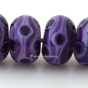 May include: Four purple glass beads with white swirls and dots. The beads are strung on a white thread.