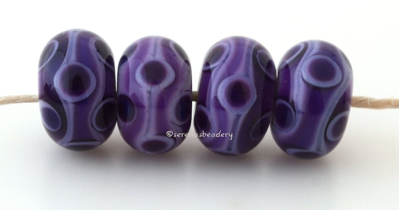 May include: Four purple glass beads with white swirls and dots. The beads are strung on a white thread.