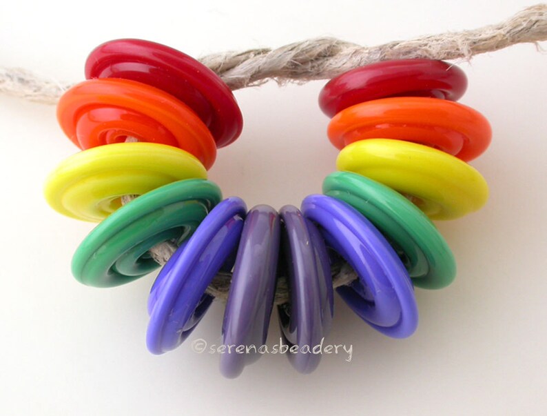 K&ouml;nnte beinhalten: Ein Regenbogen aus bunten Glasperlen, die auf einem St&uuml;ck Schnur aufgef&auml;delt sind. Die Perlen sind in der Reihenfolge der Farben des Regenbogens angeordnet, beginnend mit Rot und endend mit Lila.