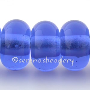 May include: Five blue glass beads with a horizontal white and dark blue stripe. The beads are strung on a natural fiber cord. The beads have a glossy finish and are oval-shaped. The text "serenasbeadery" is visible on the beads.
