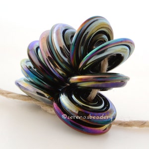 May include: A cluster of iridescent black glass beads, each with a ring shape, are strung together on a natural twine cord. The beads are arranged in a spiral pattern, creating a unique and eye-catching design. The beads have a metallic sheen and reflect light in a rainbow of colors.