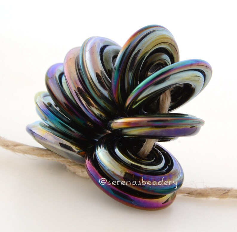 May include: A cluster of iridescent black glass beads, each with a ring shape, are strung together on a natural twine cord. The beads are arranged in a spiral pattern, creating a unique and eye-catching design. The beads have a metallic sheen and reflect light in a rainbow of colors.