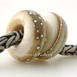 May include: Two white glass beads with silver accents. The bead on the left has a brown swirl and silver dots. The bead on the right has silver lines. The beads are on a silver chain.
