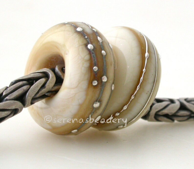 May include: Two white glass beads with silver accents. The bead on the left has a brown swirl and silver dots. The bead on the right has silver lines. The beads are on a silver chain.