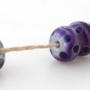 May include: Four purple and white glass beads with a swirling pattern are strung on a natural twine thread. The beads are round and have a glossy finish.