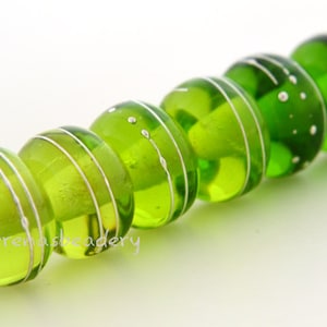 May include: A row of five translucent lime green glass beads strung on a light-colored cord. Each bead features thin, horizontal silver-colored wire accents wrapped around the circumference, with some beads displaying small silver-colored dots.